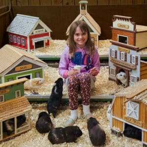little girl sitting on a step smiling with miniature houses and guinea pigs around her with a cup of lettuce in her hand